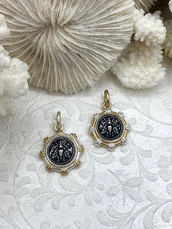 May include: Two gold and silver bee charms with black enamel. The bee charms are round and have a detailed design. The charms are on a white background with a floral pattern.