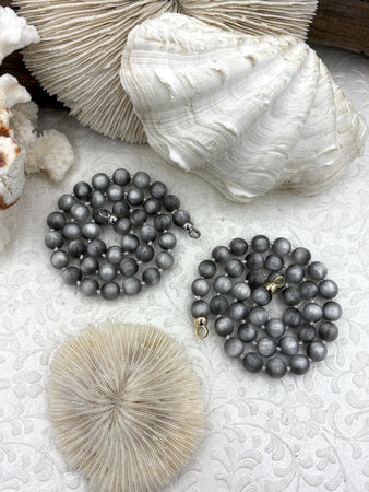 May include: Two strands of round, dark gray beads arranged in circular coils. One strand features a silver-toned metal clasp, while the other strand is finished with a gold-toned metal clasp. The beads have a smooth, uniform, and slightly reflective surface.